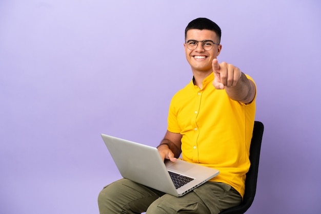 Hombre joven sentado en una silla con portátil le señala con el dedo con una expresión de confianza