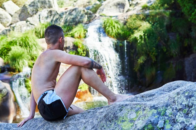 Hombre joven sentado sobre una roca en bañador, relajado en un río con cascada.