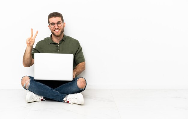 Foto hombre joven sentado en el suelo sonriendo y mostrando el signo de la victoria