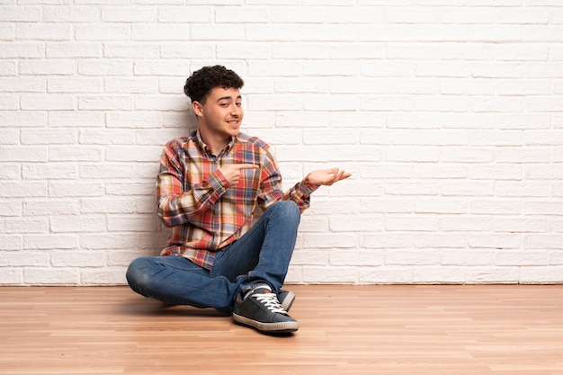 Hombre joven sentado en el suelo sosteniendo copyspace imaginario en la palma para insertar un anuncio