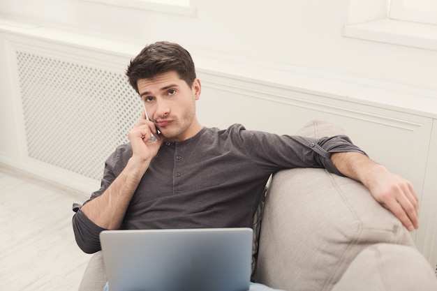 Hombre joven serio con gadgets. Freelancer consultando en el móvil mientras trabaja en una computadora portátil, copie el espacio