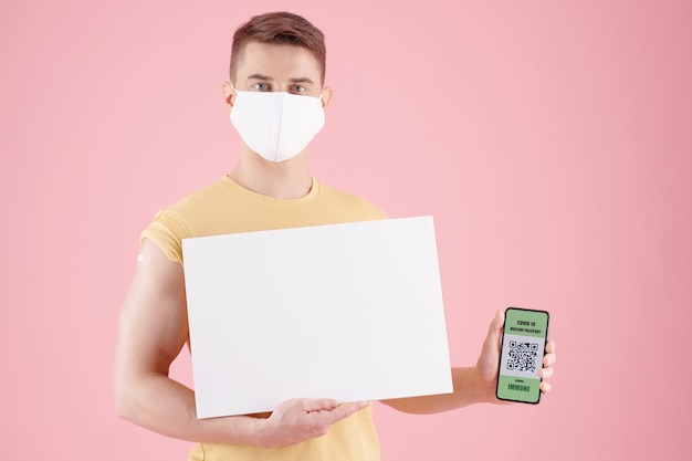 Hombre joven serio en máscara protectora que muestra cartel negro y teléfono inteligente con código qr y p ...