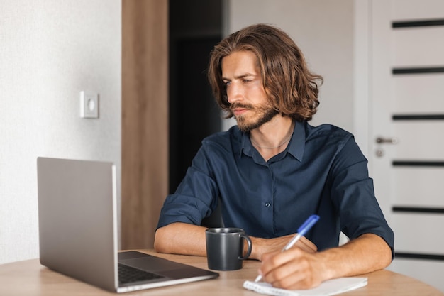 El hombre joven se sienta a la mesa y escribe en un cuaderno mientras mira en una computadora portátil