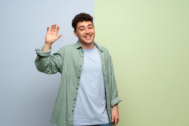 Hombre joven sobre fondo azul y verde que saluda con la mano con la expresión feliz