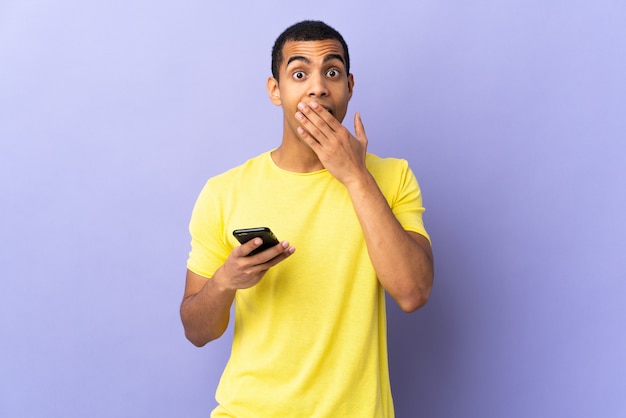 Hombre joven sobre pared púrpura aislada usando teléfono móvil con expresión facial sorpresa