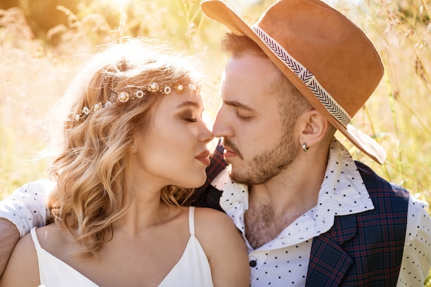 Un hombre joven con un sombrero y una hermosa niña con maquillaje y peinado, abrazando sentado en un campo en la hierba en un día soleado