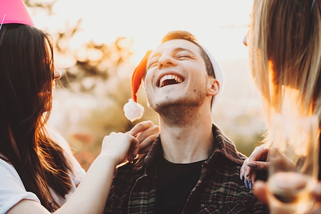 Foto hombre joven con sombrero de navidad manteniendo los ojos cerrados y riendo con dos mujeres encantadoras.