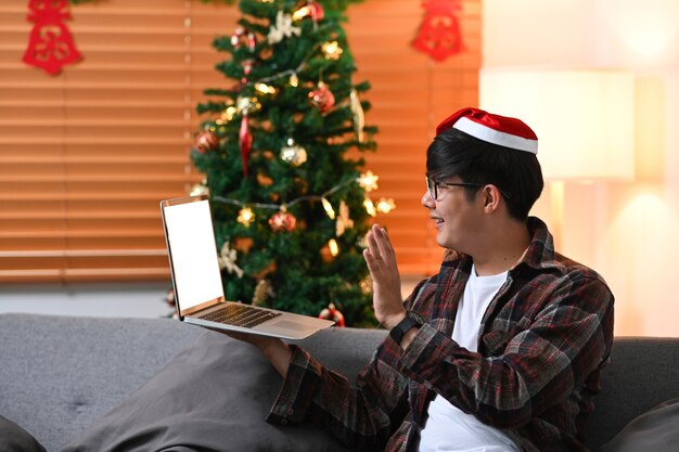 Hombre joven con sombrero rojo de Santa con videollamada en la computadora portátil con su familia en Navidad.