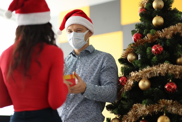 Hombre joven con sombrero de santa claus y máscara protectora en la cara dando regalo a la mujer cerca del árbol de navidad