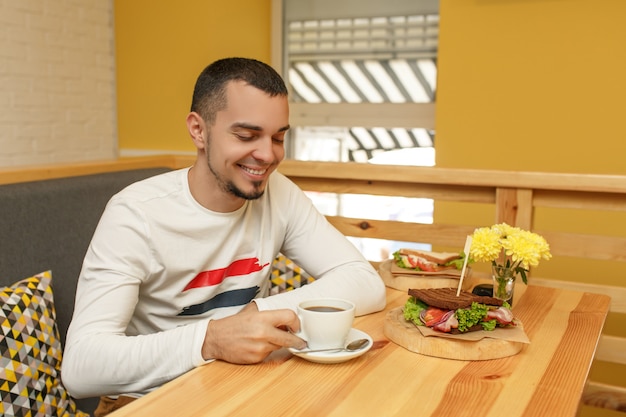 El hombre joven sonríe y toma la taza con café, desayuno en café