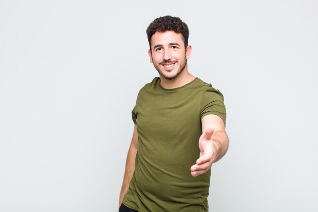 Hombre joven sonriendo, luciendo feliz, confiado y amigable, ofreciendo un apretón de manos para cerrar un trato, cooperando