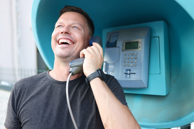 Hombre joven sonriente hablando por cabina telefónica en la calle. Comunicación por concepto de teléfono fijo