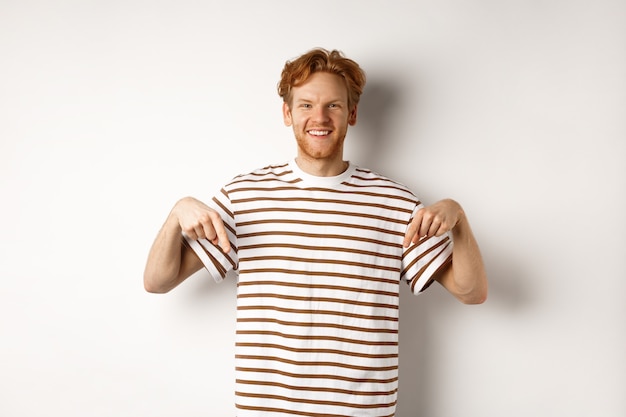 Hombre joven sonriente con el pelo rojo desordenado y la barba, señalando con el dedo hacia abajo, recomendando la tienda, de pie sobre fondo blanco.