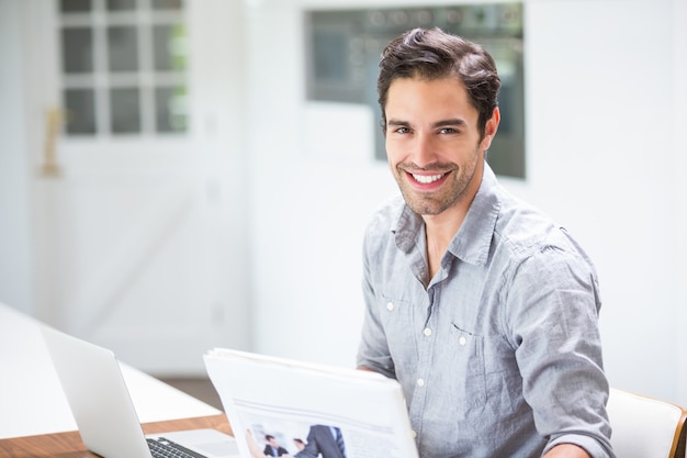 Foto hombre joven sonriente que lleva a cabo documentos mientras que se sienta en el escritorio con la computadora portátil