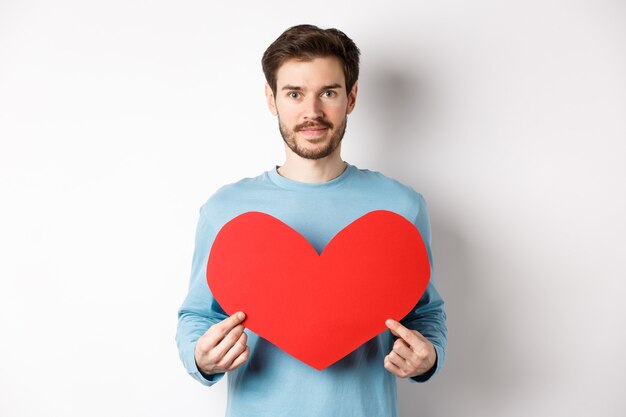 Hombre joven sonriente que sostiene el recorte del corazón de San Valentín, esperando el verdadero amor de la novia, de pie en blanco