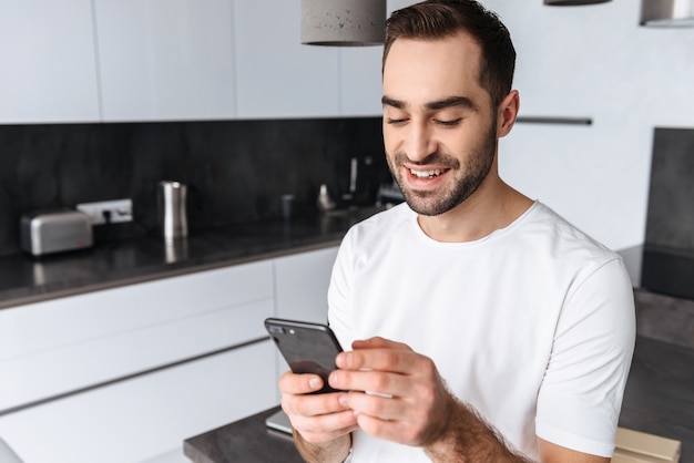 Hombre joven sonriente que sostiene el teléfono móvil mientras está de pie en la cocina de casa