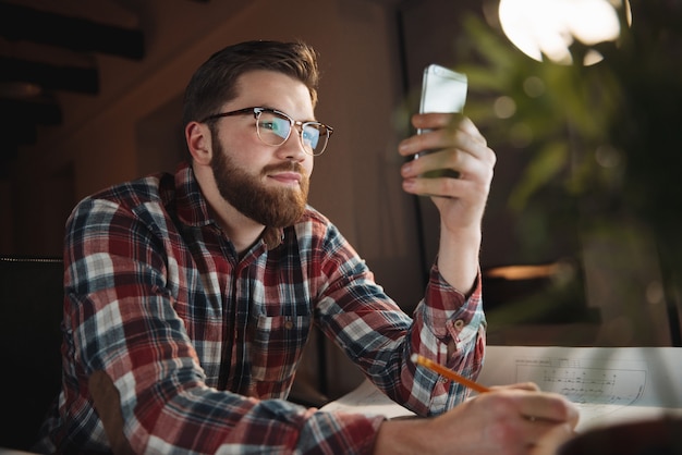 Hombre joven sonriente que usa su teléfono inteligente mientras está sentado en su lugar de trabajo