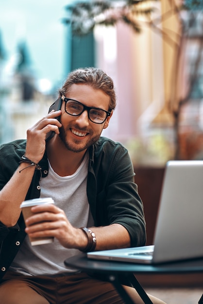 Hombre joven sonriente en ropa casual hablando por el teléfono inteligente y usando la computadora portátil mientras trabajaba en la cafetería