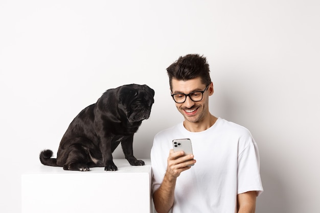 Hombre joven sonriente con smartphone y sentado cerca de perro. Propietario de Pug comprobar fotos en el teléfono móvil, fondo blanco.