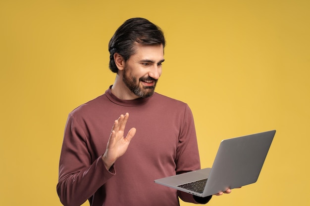 Hombre joven sonriente sosteniendo una computadora portátil mientras tiene una llamada en línea posando en un estudio aislado en amarillo