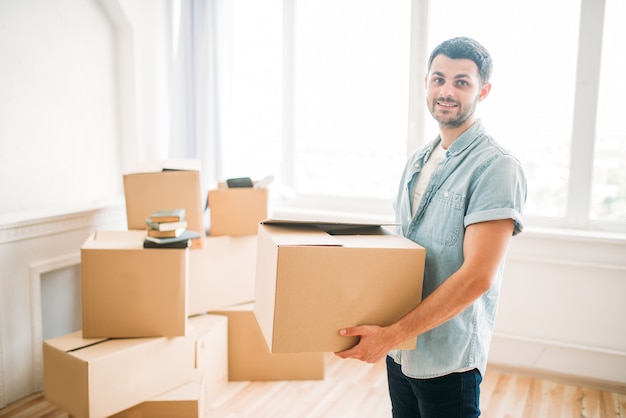 Hombre joven sonriente tiene caja de cartón en las manos, inauguración de la casa. Mudarse a una nueva casa