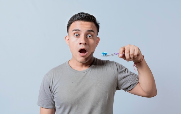 Hombre joven sorprendido sosteniendo un cepillo de dientes con pasta de dientes aislado chico con expresión wow sosteniendo un cepillo de dientes aislado en blanco Joven sorprendido sosteniendo un cepillo de dientes con pasta de dientes