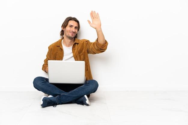 Hombre joven con su computadora portátil sentada en el suelo saludando con la mano con expresión feliz