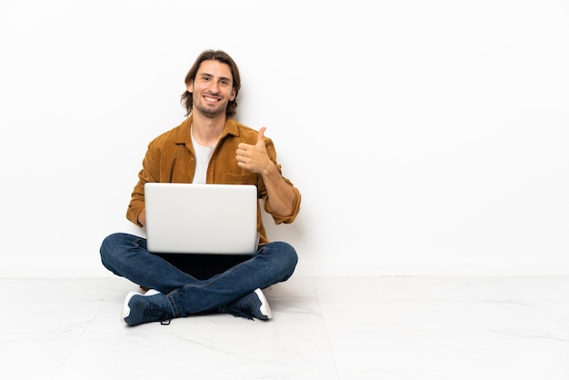 Foto hombre joven con su computadora portátil sentado en el suelo con los pulgares hacia arriba porque algo bueno ha sucedido