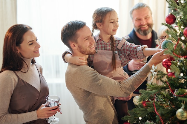 Hombre joven con su pequeña hija mirando una de las bolas de juguete decorativas en la rama del árbol de Navidad en casa