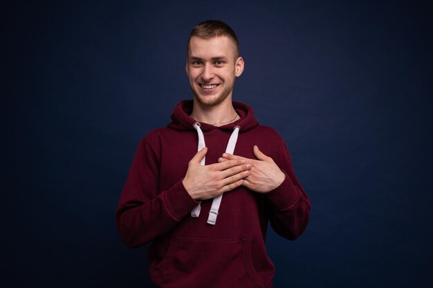 Hombre joven con una sudadera con capucha roja y jeans posando sobre un fondo azul pone sus manos a su corazón