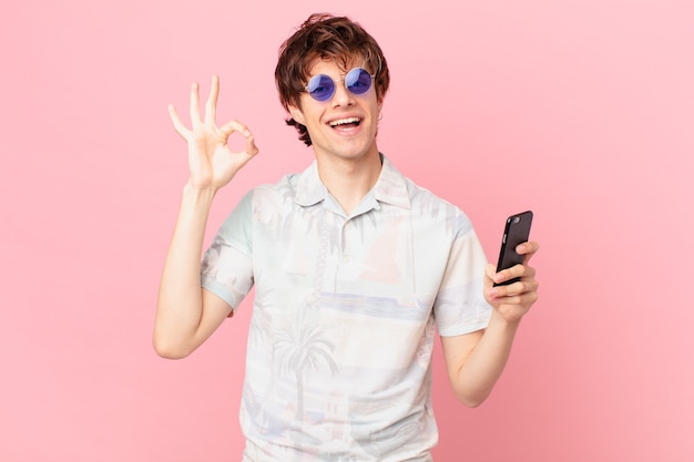 Hombre joven con un teléfono celular que se siente feliz mostrando aprobación con gesto bien