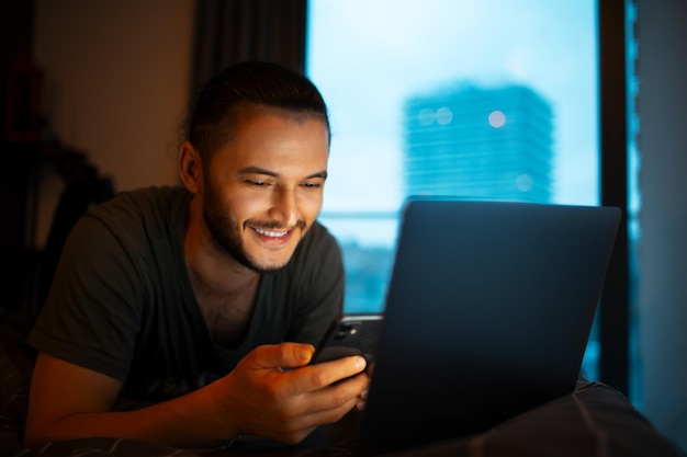 Foto hombre joven tendido en la cama usando teléfono inteligente y portátil foto oscura de la noche