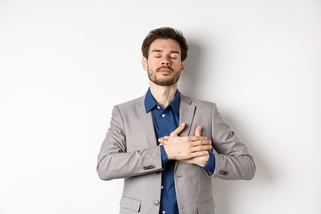 Hombre joven en traje cierra los ojos, tomados de la mano en el corazón y recuerda recuerdos felices, sintiéndose nostálgico, de pie contra el fondo blanco.