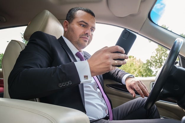 Hombre joven en traje de negocios está conduciendo un coche caro y charlando en el teléfono inteligente