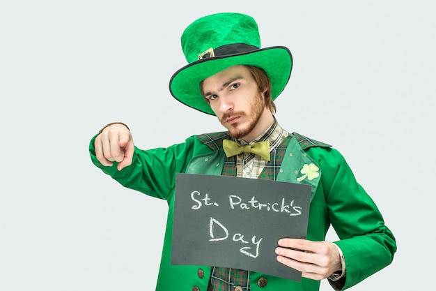 Hombre joven en traje verde con tableta oscura con la escritura de San Patricio en él. Apunta directamente y mira a la cámara. Aislado sobre fondo gris