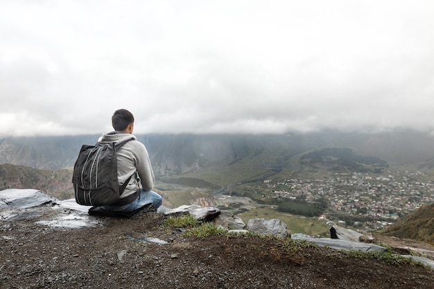 Hombre joven turista con mochila se sienta en una pendiente con el telón de fondo de las montañas del Cáucaso, Georgia en un día brumoso