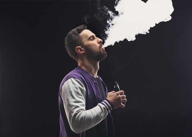 Hombre joven vaping, tiro del estudio. Chico barbudo que sopla una nube de humo sobre fondo negro. Concepto de vapor y fumar sin nicotina, espacio de copia.