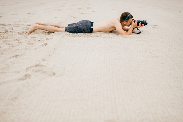 Hombre joven viajero acostado en la playa cerca del mar de vacaciones y hacer fotos de la naturaleza. Topless apuesto hombre descalzo con cámara y gran lente espiando a la gente desde lejos. Hora de pasatiempo. Niño descansando en la arena