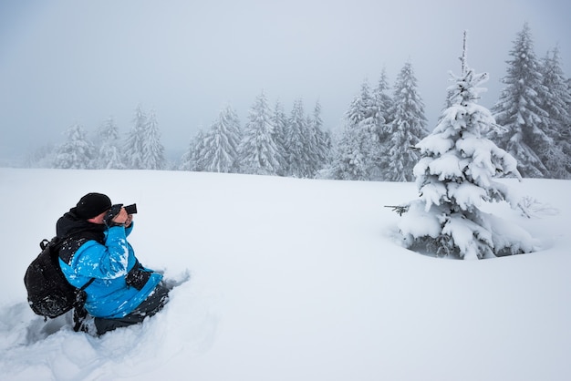 Hombre joven viajero con mochila toma fotografías de un hermoso abeto nevado alto en un gran ventisquero contra el telón de fondo de la niebla en un día de invierno helado