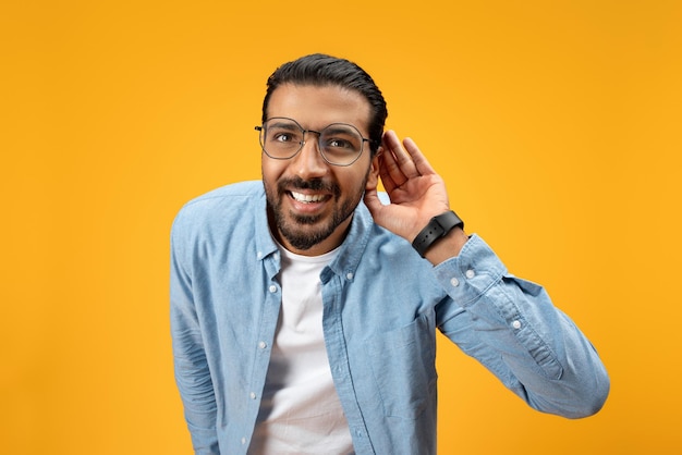 Un hombre jovial con barba y gafas se inclina con una mano a su oreja retratando una escucha o