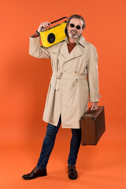 Foto hombre jubilado alegre sosteniendo boombox amarillo y maletín en manos sobre fondo naranja