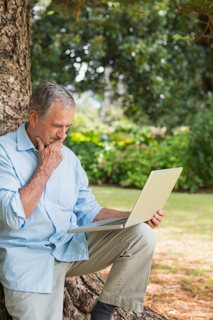 Hombre jubilado apoyado contra un árbol con una computadora portátil