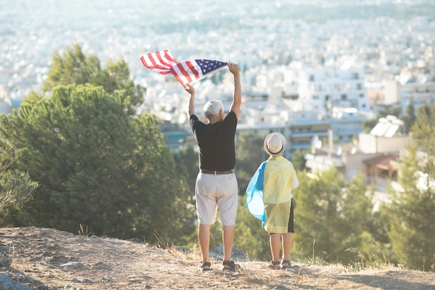 Hombre jubilado y niño sosteniendo las banderas de Estados Unidos y Ucrania a la vista de la ciudad