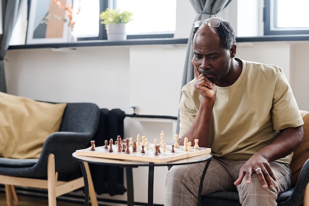 Hombre jubilado pensativo de etnia africana mirando el tablero de ajedrez en una mesa pequeña