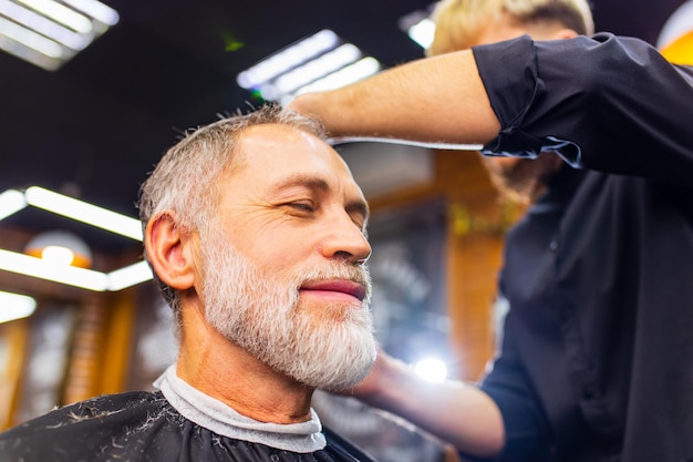 Hombre jubilado visitando la barbería sentado en la silla del peluquero mientras se hace un peinado genial