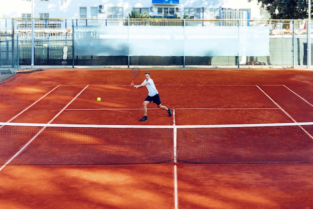 El hombre juega al tenis en el campo de tenis de arcilla vista desde lejos