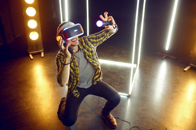 Hombre jugando en casco de realidad virtual y gamepad en cubo luminoso. Interior oscuro del club de juego, tecnología VR con visión 3D