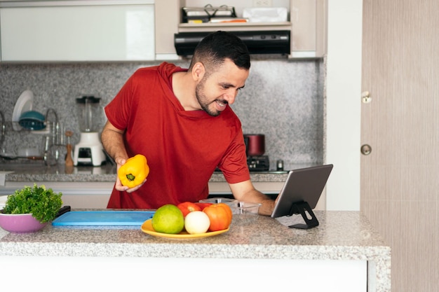 Hombre latino consultando una receta en línea con una tableta mientras prepara una comida en la cocina de su casa