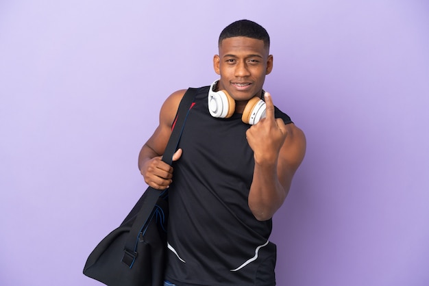 Hombre latino de deporte joven con bolsa de deporte aislado en púrpura haciendo gesto que viene