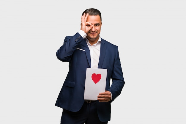 El hombre latino envejecido medio que celebra el día de tarjetas del día de San Valentín confiado haciendo gesto aceptable en ojo